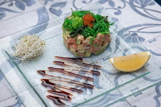 Elegant salmon and avocado tartare with seaweed and sprouts on a glass plate, garnished with lemon and sauce.